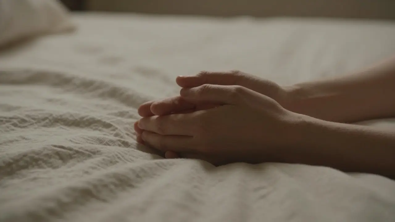 Two hands gently holding each other on a bedsheet, symbolizing quiet connection and emotional closeness.